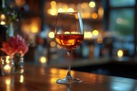 Glass of amber wine on wooden table with warm bokeh lights and red flower in soft focus background, suitable for restaurant, bar, and beverage marketing.