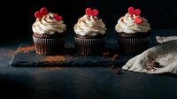 Three chocolate cupcakes with cream frosting and red heart decorations on dark slate board for Valentine's Day celebration and romantic dessert occasions.