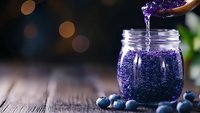 Fresh blueberry jam dripping from wooden spoon into glass jar on rustic wooden table with fresh berries and soft bokeh background. Food photography.
