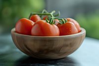 Fresh ripe tomatoes on vine in wooden bowl against blurred green background, close-up view with soft natural lighting and shallow depth of field.