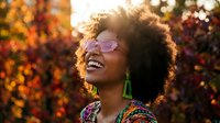Happy young African American woman with curly afro hair and glasses laughing joyfully outdoors in warm autumn sunlight with colorful fall foliage background.