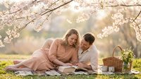 Couple reading together under blooming cherry blossom trees in spring garden, romantic picnic scene with basket and flowers for lifestyle and relationship content.