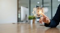 Businessman in dark suit touching glowing gear icons floating above wooden desk in modern office, representing technology, innovation, and digital transformation for business solutions.