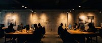 Silhouettes of diverse professionals working at wooden desks in a modern office with warm ambient lighting, suitable for corporate, business, and workplace concepts.