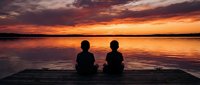 Two people sitting on wooden dock watching dramatic sunset over calm lake with orange and purple sky reflections in water creating peaceful moment.