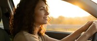 Young woman with long curly dark hair and olive complexion driving car during golden hour sunset, hands on steering wheel, looking ahead with serene expression, natural lighting through windshield.