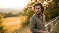 Handsome middle-aged Caucasian man with wavy hair and beard smiling outdoors during golden hour sunset in rural countryside landscape.