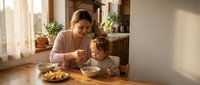 Adult woman with medium skin tone and brown hair, wearing casual beige clothing, feeding toddler boy at wooden table during mealtime in bright home kitchen setting.