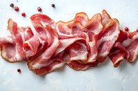 Thin slices of raw bacon arranged in a row on white textured background with peppercorns and red berries for gourmet food photography.