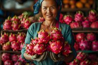 Asian female vendor holding fresh dragon fruit at traditional market with colorful produce display in background.