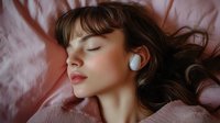 Young woman with closed eyes wearing wireless earbuds lying on pink silk bedding, dark wavy hair spread out, peaceful relaxed expression, listening to music.