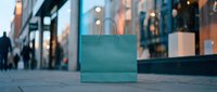 Teal shopping bag on urban street with blurred storefronts and pedestrians in background, retail and commerce concept for marketing.