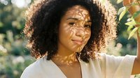 Young African American woman with curly hair enjoying dappled sunlight through leaves creating natural shadow patterns on her face in outdoor garden setting.