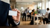 Businessman in dark suit holding blank white business card in modern office environment with colleagues working in background, networking and corporate communication concept.