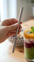 Hand holding stainless steel reusable drinking straw next to layered smoothie beverage with mint leaf garnish on wooden table, eco-friendly sustainable lifestyle.