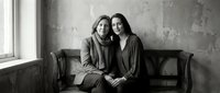 Two women sitting together on vintage leather sofa in minimalist interior with concrete wall. Black and white portrait of female friends embracing indoors.