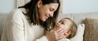 Happy young mother in cream sweater tenderly holding and gazing at her smiling baby while sitting on beige sofa in cozy living room during bonding moment.