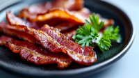 Crispy fried bacon strips on black plate garnished with fresh parsley. Close-up view shows texture and caramelization of cooked meat for food photography.