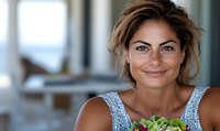 Smiling Mediterranean woman with tousled hair holding fresh salad greens, wearing casual summer top. Natural daylight setting creates warm ambiance.