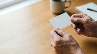 Mature man with medium skin tone holding blank white card at wooden desk with coffee mug and notebook, suitable for business, branding, and corporate messaging concepts.