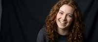 Young Caucasian woman with curly red hair smiling warmly in gray sweater against dark background for professional headshot and portrait photography.