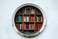 Round window with wooden bookshelves filled with colorful books against white wall, creating unique architectural library display for reading room design.