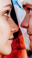Close-up profile view of young couple facing each other with tender expressions against vibrant orange and blue background, capturing intimate romantic moment.