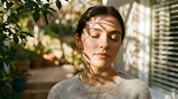 Young Caucasian woman with closed eyes enjoying warm sunlight streaming through window blinds creating shadow patterns on face in cozy indoor setting.