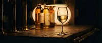 Glass of white wine on wooden table with warm ambient lighting and bottles in background creating cozy atmosphere for dining and entertainment.