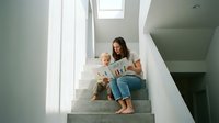 Young mother reading book with toddler son on modern home staircase in bright natural light creating warm family bonding moment for parenting content.