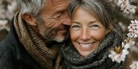 Happy mature Caucasian couple embracing outdoors in spring with cherry blossoms, wearing cozy knit scarves, showing love and togetherness in nature.