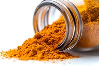 Ground turmeric powder spilling from glass jar on white surface, vibrant orange-yellow spice with selective focus showing texture and aromatic detail.