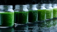 Fresh green smoothie or juice in glass jars arranged in row on dark reflective surface with water drops, showing healthy beverage preparation.
