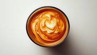 Creamy caramel latte with heart-shaped foam art in ceramic cup on neutral background, viewed from above.