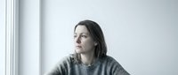 Thoughtful young Caucasian woman in gray sweater sitting by window with natural light, contemplating future plans and personal growth opportunities.