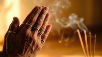 Hands pressed together in prayer pose with rings, incense smoke rising softly in warm golden light, evoking spirituality and mindfulness.