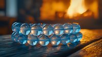 Translucent blue glass beads bracelet on wooden surface with warm fireplace glow in background creating atmospheric mood lighting and reflections.