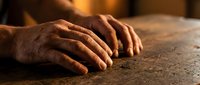 Elderly hands resting on wooden table surface in warm golden light showing age and wisdom through weathered skin texture and gentle positioning.