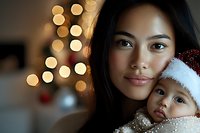 Young Asian mother holding baby in Santa hat against Christmas lights background, creating intimate holiday moment with soft bokeh glow and warm atmosphere.