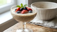 Chia seed pudding topped with fresh strawberries and blueberries in a coupe glass, served with a ceramic bowl on a wooden surface near a window.