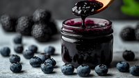 Homemade blackberry jam being spooned from glass jar surrounded by fresh blueberries and blackberries on rustic wooden surface for breakfast spreads.