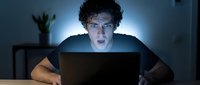 Young man with curly dark hair and light skin tone, wearing casual tan shirt, working late at night on laptop with focused expression, illuminated by screen glow in dark office environment.