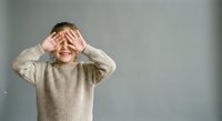 Young boy in beige knit sweater covering eyes with hands in playful peek-a-boo gesture against neutral gray background. Child expressing joy and innocence.