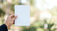 Person holding blank white paper outdoors with blurred green foliage background, suitable for mockups, presentations, and advertising campaigns.