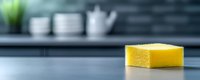 Yellow kitchen sponge on clean counter with blurred modern kitchen background featuring white dishes and plant for household cleaning concepts.