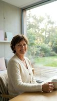 Mature woman with short brown hair and Caucasian ethnicity sitting at wooden desk by large window, holding mug, wearing cream cardigan, smiling at camera with natural daylight and garden view.