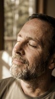 Middle-aged man with gray beard and receding hairline in profile, eyes closed in peaceful expression, indoor setting with window light, contemplative mood for wellness and mindfulness concepts.