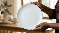 Cropped view of male hands holding empty white plate in kitchen setting with natural light and blurred background for food service or dining concepts.