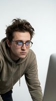 Young Caucasian man with messy brown hair wearing glasses and olive hoodie working on laptop computer against white background for remote work concepts.