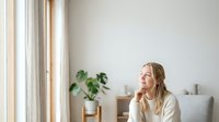 Woman in casual cream sweater sitting by window, looking thoughtful and contemplative in bright, minimalist interior space with natural light and potted plant.